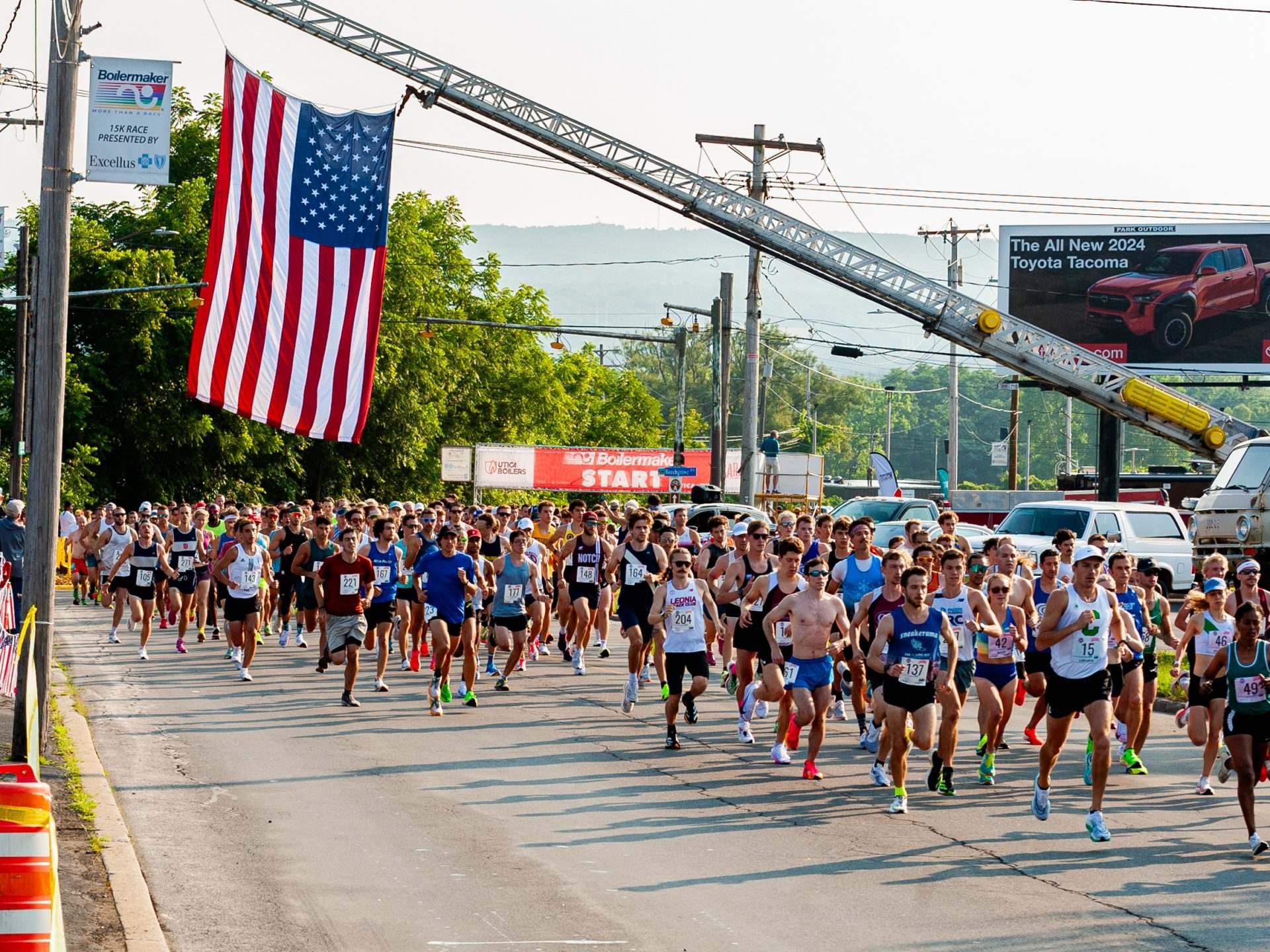 A group of runners just after the 15K start line run beneath a large American flag.