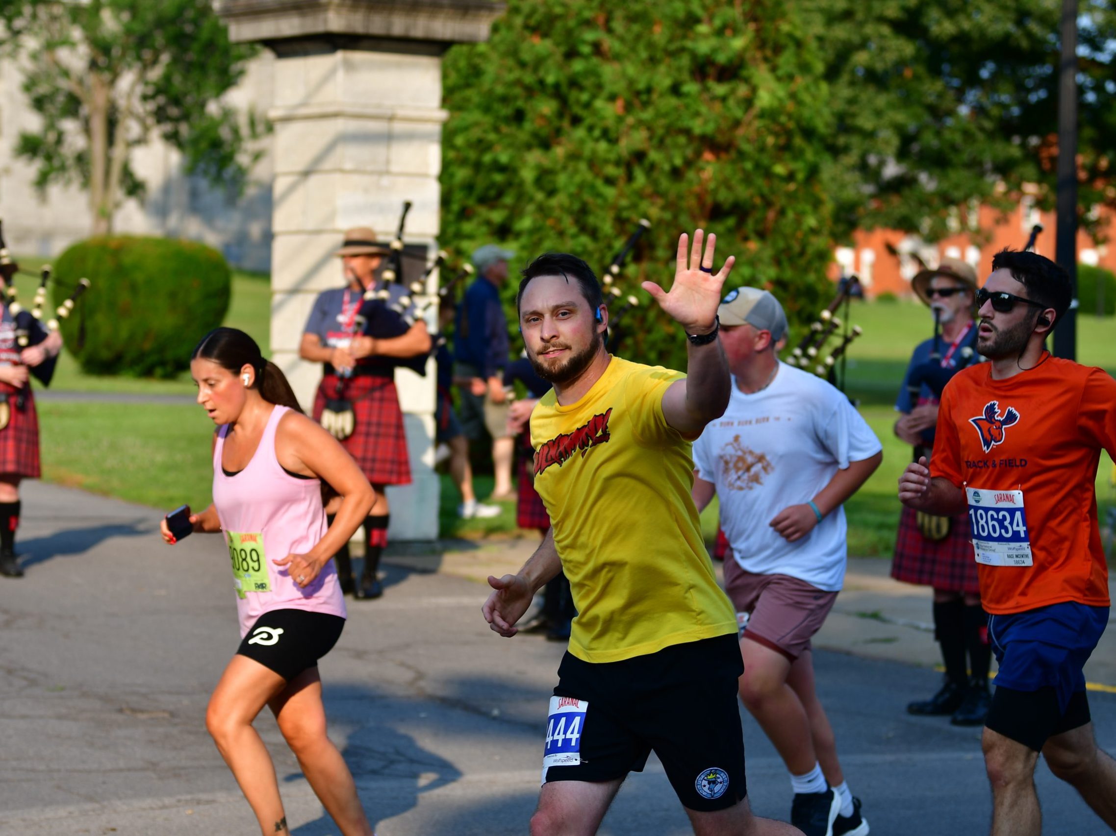A man in a yellow shirt waves at the camera along the 5K course with bagpipes in the background playing