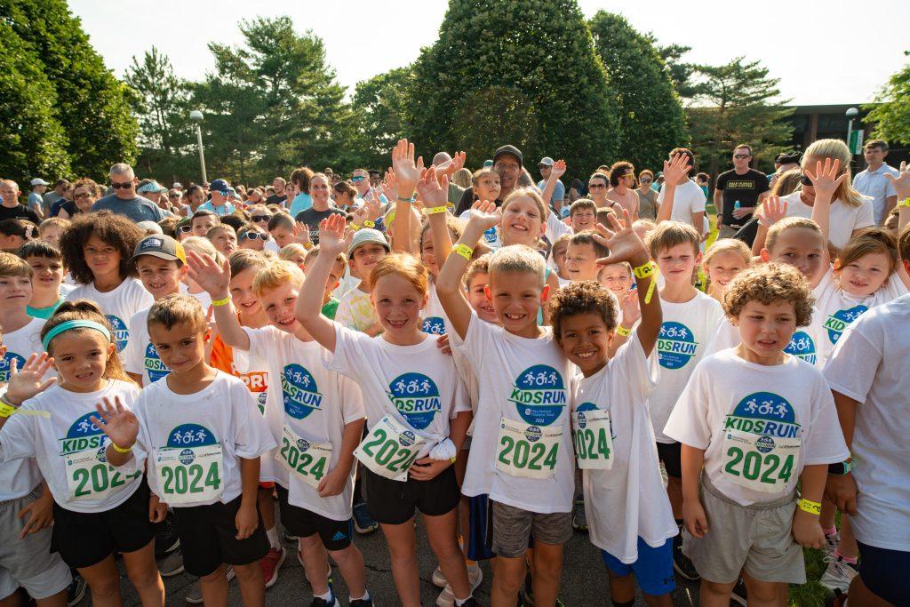 A group of kids grin and raise their hands in their matching shirts at the Kids Run